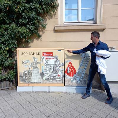 Angestellter vom Stadtarchiv zeigt auf einen Stromkasten mit künstlerischem Bild